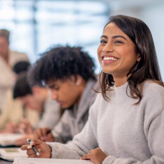 A group of multi-ethnic post secondary students sit in class with their textbooks open in front of them as they each individually study.  The focus is on a girl of Indian decent who is looking up from her studies and smiling.